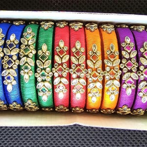 May include: A set of 15 colorful silk thread bangles with intricate designs and embellishments. The bangles are arranged in a box and feature a variety of colors, including blue, green, red, yellow, and purple.