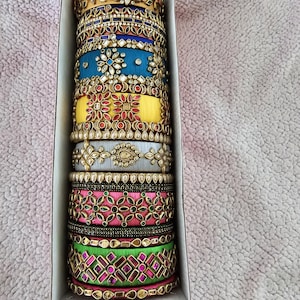 May include: A stack of colorful silk thread bangles with intricate gold and jeweled designs. The bangles are arranged in a box, with the top bangle featuring a floral design.
