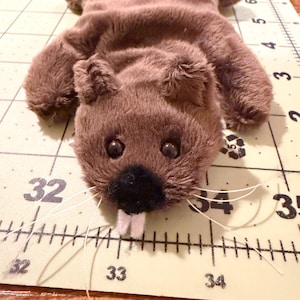 May include: A plush brown beaver toy with a black nose, white whiskers, and two white teeth. The toy has small black eyes and is lying on a cutting mat with measurements in inches.