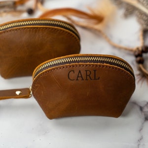 May include: Two brown leather zippered pouches with a gold zipper. The smaller pouch has the name "CARL" engraved on the front.