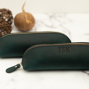 May include: Two dark green leather pencil cases with gold zippers. The smaller case has the initials "YSK" embossed on the front.