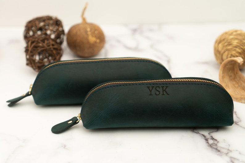 May include: Two dark green leather pencil cases with gold zippers. The smaller case has the initials "YSK" embossed on the front.