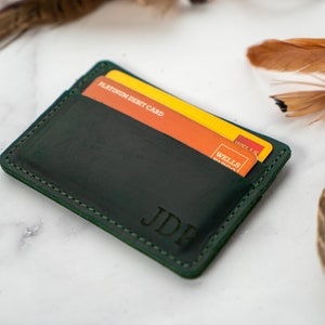 May include: A dark green leather cardholder with two credit cards visible. The cardholder has the initials "JDE" embossed on the front. The stitching is a lighter green. The background includes feathers.
