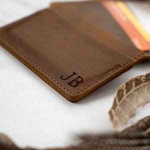May include: A brown leather wallet with the initials "JB" engraved on the front. The wallet is open, revealing a few cards. The edges are stitched with a contrasting thread. Decorative feathers are in the background.