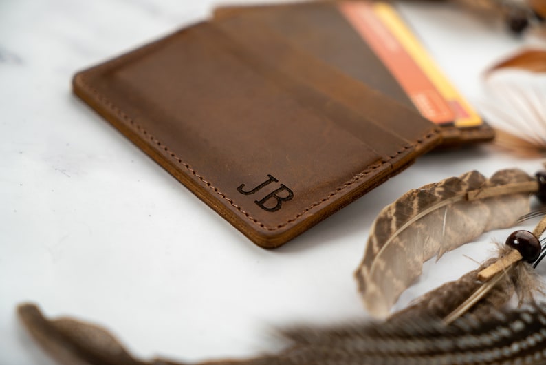 May include: A brown leather wallet with the initials "JB" engraved on the front. The wallet is open, revealing a few cards. The edges are stitched with a contrasting thread. Decorative feathers are in the background.