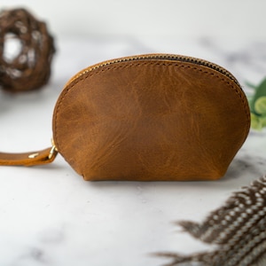 May include: A small, brown leather pouch with a zipper closure and a strap. The pouch is made of a soft, textured leather and has a slightly curved shape.