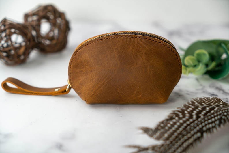 May include: A small, brown leather pouch with a zipper closure and a strap. The pouch is made of a soft, textured leather and has a slightly curved shape.