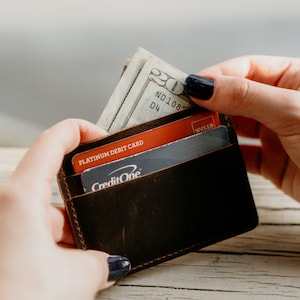 May include: A dark brown leather wallet with a few credit cards and a twenty-dollar bill. The wallet is held open, revealing the contents. The stitching is visible around the edges of the wallet.
