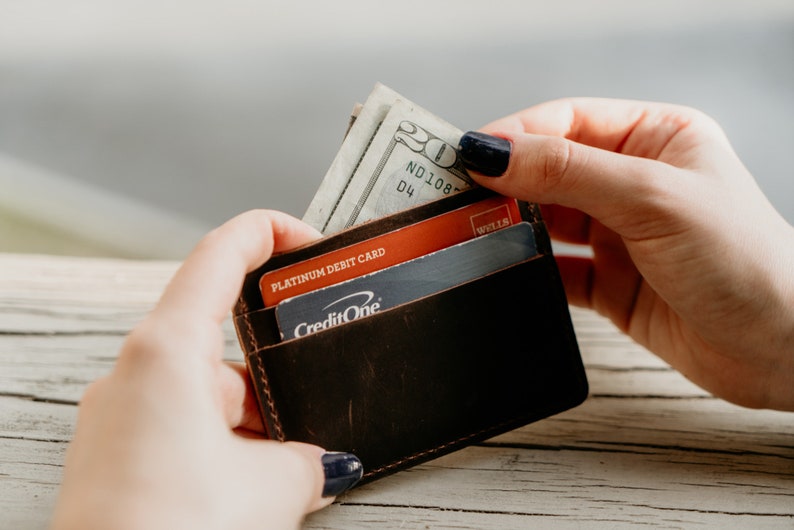 May include: A dark brown leather wallet with a few credit cards and a twenty-dollar bill. The wallet is held open, revealing the contents. The stitching is visible around the edges of the wallet.