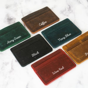 May include: A collection of leather cardholders in various colors: army green, coffee, navy blue, black, brown, and wine red. Each cardholder has a minimalist design with visible stitching and a slot for cards. The names of the colors are written in white script.