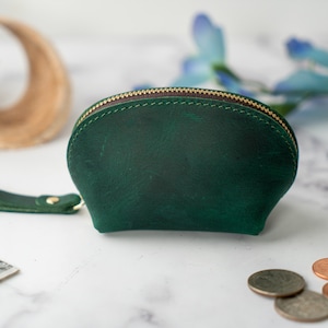 May include: A small, green leather coin purse with a zipper closure and a strap. The purse is on a white marble surface.