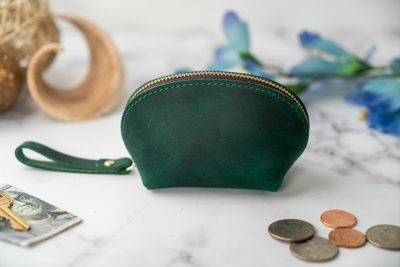 May include: A small, green leather coin purse with a zipper closure and a strap. The purse is on a white marble surface.