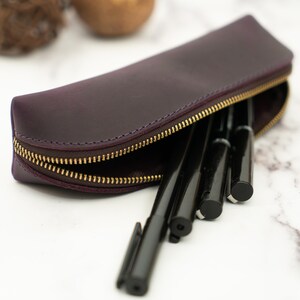 K&ouml;nnte beinhalten: Eine violette Lederstiftetui mit einem goldenen Rei&szlig;verschluss. Das Etui ist ge&ouml;ffnet und mit schwarzen Stiften gef&uuml;llt.