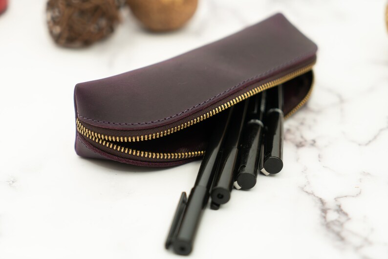 K&ouml;nnte beinhalten: Eine violette Lederstiftetui mit einem goldenen Rei&szlig;verschluss. Das Etui ist ge&ouml;ffnet und mit schwarzen Stiften gef&uuml;llt.