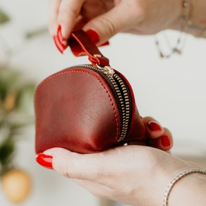 May include: A small, red leather coin purse with a gold zipper. The purse is being held open by a hand with red nail polish.