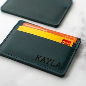 May include: Teal leather card holder with the name "KAYLA" engraved on the front. The card holder contains a yellow and orange credit card. The card holder is rectangular and has stitched edges.