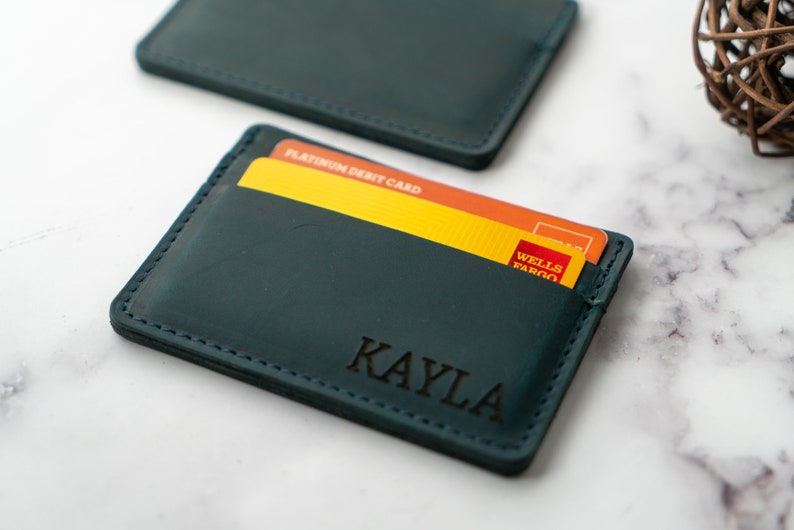 May include: Teal leather card holder with the name "KAYLA" engraved on the front. The card holder contains a yellow and orange credit card. The card holder is rectangular and has stitched edges.