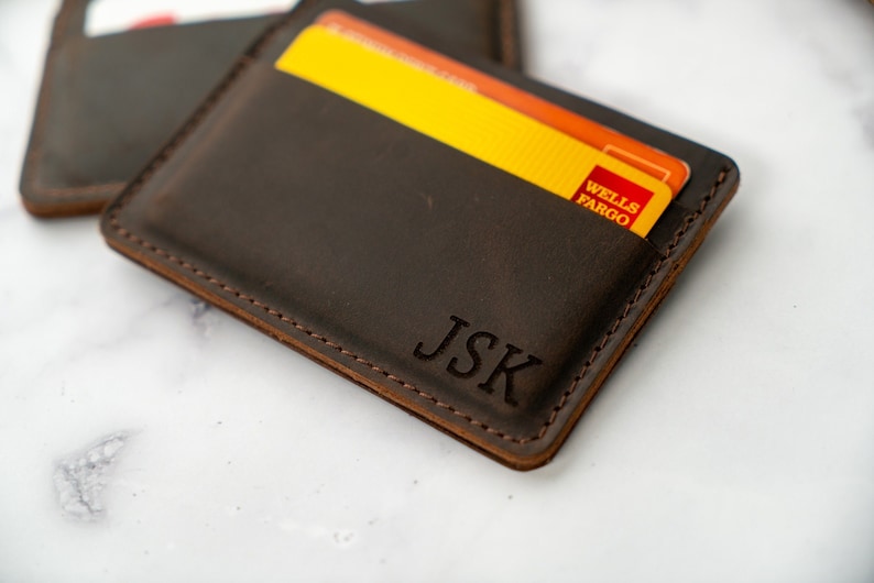 May include: A dark brown leather cardholder with the initials "JSK" embossed on the front. The cardholder has a stitched border and contains a yellow and orange Wells Fargo credit card. Another cardholder is visible in the background.