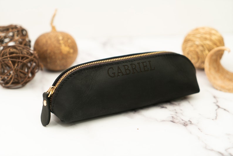 May include: Black leather pencil case with a gold zipper and the name "GABRIEL" embossed on the front.