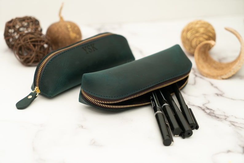 May include: Two green leather pencil cases with gold zippers. One case has the initials "YSK" embossed on the front. The other case is open and has black pens inside.