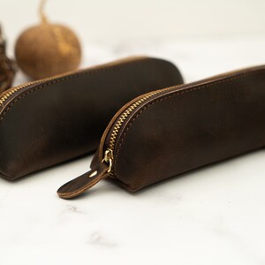 May include: Two brown leather pencil cases with gold zippers. The cases are lying on a white marble surface.