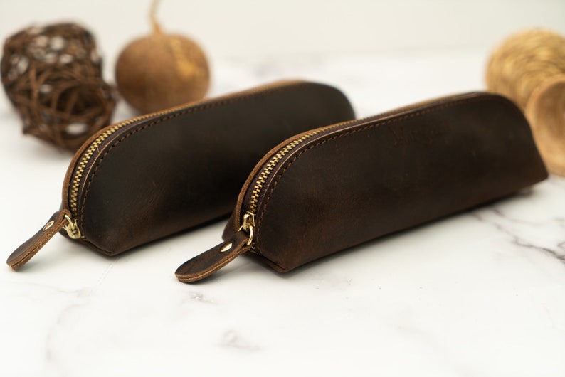 May include: Two brown leather pencil cases with gold zippers. The cases are lying on a white marble surface.