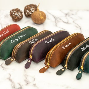 May include: Seven leather pencil cases in different colors, including wine red, army green, coffee, purple, brown, black, and navy blue. Each case has a gold zipper and is labeled with its color.