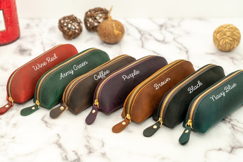 May include: Seven leather pencil cases in different colors, including wine red, army green, coffee, purple, brown, black, and navy blue. Each case has a gold zipper and is labeled with its color.
