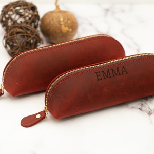 May include: Two red leather pencil cases with gold zippers. The smaller case is plain, while the larger case has the name "EMMA" engraved on it.