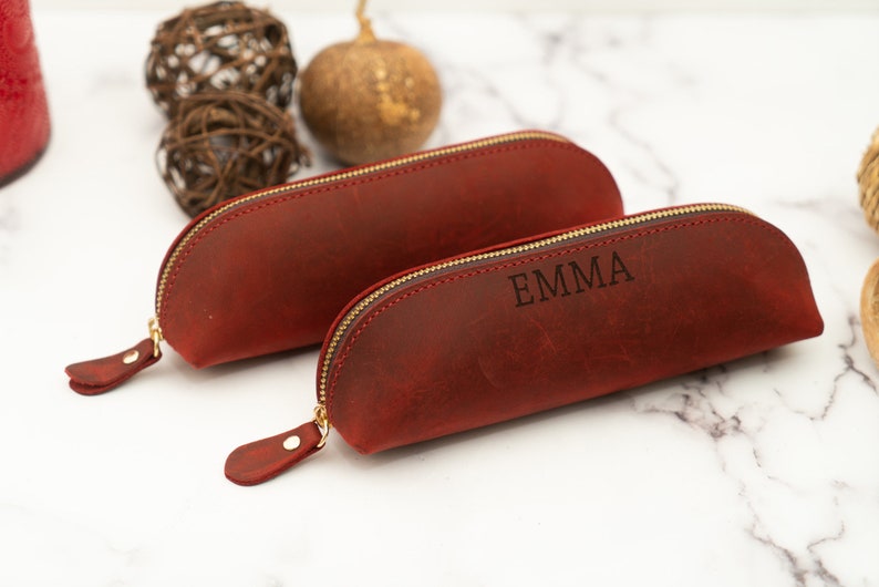 May include: Two red leather pencil cases with gold zippers. The smaller case is plain, while the larger case has the name "EMMA" engraved on it.