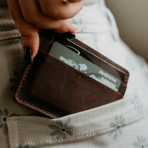 May include: A brown leather wallet with a card inside is being placed into a pocket. The wallet is a dark brown color and appears to be made of high-quality leather. The card has the American Express logo.