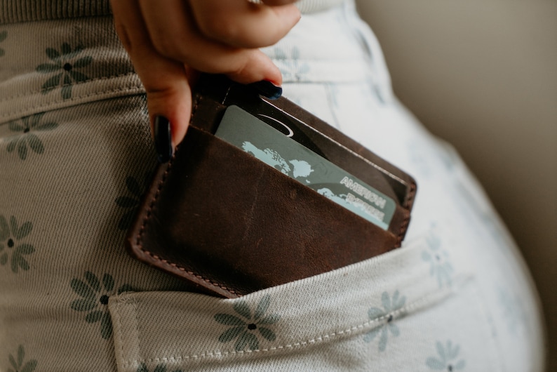 May include: A brown leather wallet with a card inside is being placed into a pocket. The wallet is a dark brown color and appears to be made of high-quality leather. The card has the American Express logo.