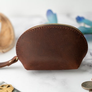 May include: A brown leather coin purse with a zipper closure and a small strap. The purse is on a white marble surface.