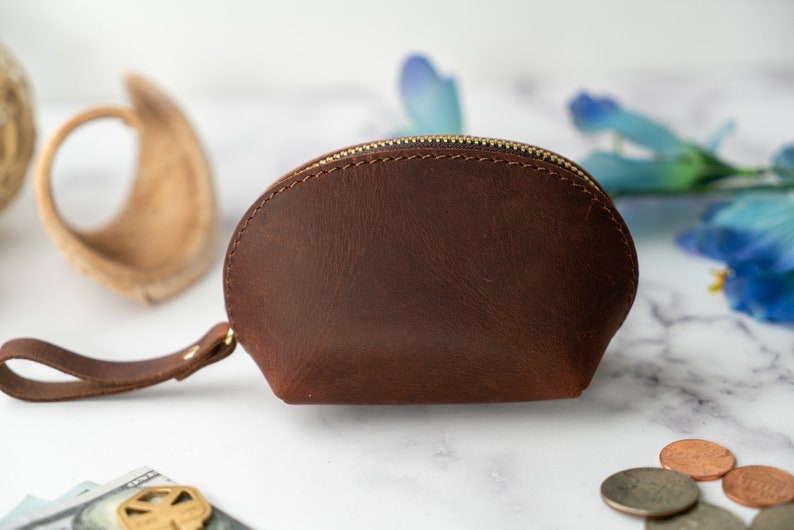 May include: A brown leather coin purse with a zipper closure and a small strap. The purse is on a white marble surface.