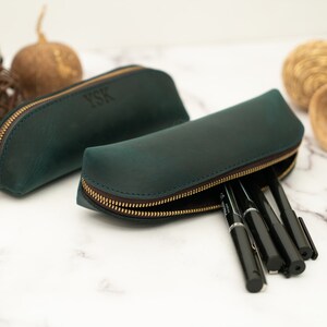 K&ouml;nnte beinhalten: Zwei gr&uuml;ne Bleistiftetuis aus Leder mit goldenen Rei&szlig;verschl&uuml;ssen. Ein Etui hat die Initialen "YSK" auf der Vorderseite gepr&auml;gt. Das andere Etui ist ge&ouml;ffnet und enth&auml;lt schwarze Stifte.