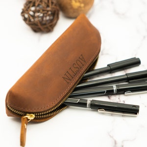 May include: A brown leather pencil case with a zipper closure. The case is personalized with the name "AUSTIN" in a simple font. The case is filled with black pens.