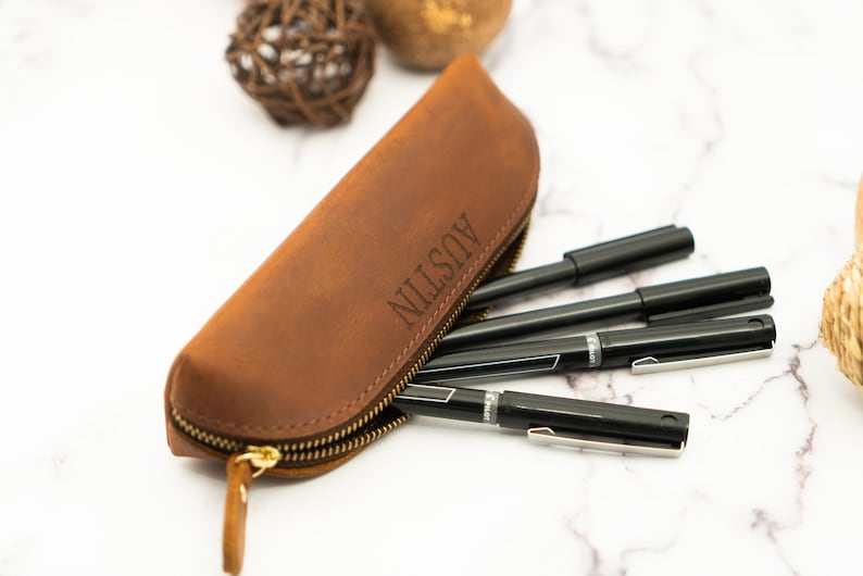 May include: A brown leather pencil case with a zipper closure. The case is personalized with the name "AUSTIN" in a simple font. The case is filled with black pens.