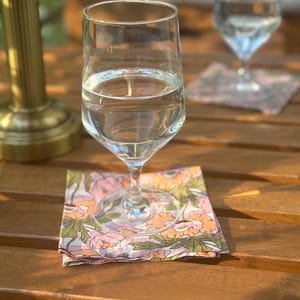 Puede incluir: Dos copas de agua de cristal transparente sobre posavasos de tela cuadrados con un estampado floral de ilustraciones botánicas en rosa, naranja y verde. Los posavasos están sobre una superficie de madera de listones junto a una lámpara de metal dorado.