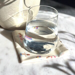 Op de afbeelding: Een helder glas water staat op een beige onderzetter met een strandscène. De onderzetter toont illustraties van mensen in badkleding, strandballen en andere zomerelementen. Op de achtergrond staat een grote, gebroken witte keramische pot.