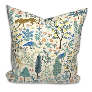 May include: Square decorative pillow with a cream-colored background featuring a detailed print of a garden scene. The design includes a leopard, peacocks, trees with yellow fruit, and various flowers in shades of blue, orange, and yellow.