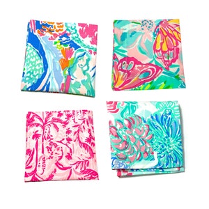 May include: Four folded fabric squares with colorful, tropical-themed patterns. The designs include mermaids, flowers, and palm trees in shades of pink, blue, green, and yellow. The squares are arranged on a white background.