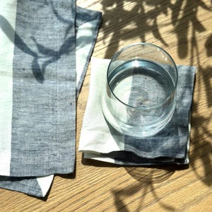 Puede incluir: Un vaso de agua transparente descansa sobre una servilleta de lino a rayas azules y blancas. Otra servilleta está doblada a la izquierda. Las servilletas y el vaso están sobre una superficie de madera, con sombras de una planta encima.