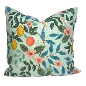 May include: Square throw pillow with a light blue background and a botanical pattern. The design features yellow lemons, orange fruit, pink flowers, and green and blue leaves. The pillow is a decorative item for a bed or sofa.