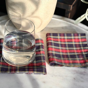 May include: A clear glass of water sits on a red, navy, and yellow plaid coaster. Two additional plaid coasters are next to the glass. The coasters are square and folded. A neutral-colored vase is in the background.