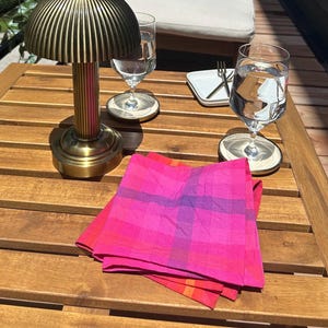 Könnte beinhalten: Eine Tischlampe aus Messing mit geripptem Schirm steht auf einem Holztisch neben zwei Weingläsern und einem Stapel rosa und rot karierter Servietten. Ein kleiner weißer Teller mit einer Gabel befindet sich ebenfalls auf dem Tisch.