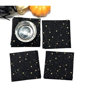 May include: Four black square cloth napkins with a gold star pattern. One napkin has a silver glass on top. The napkins are arranged on a white surface. The image has a Halloween theme.