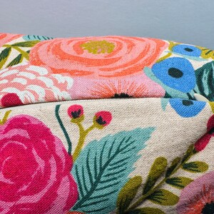 May include: Close-up of a floral patterned pillow. The pillow features large, colorful flowers in shades of pink, orange, and blue, set against a beige background. Green leaves and stems complete the design, creating a vibrant, botanical theme.