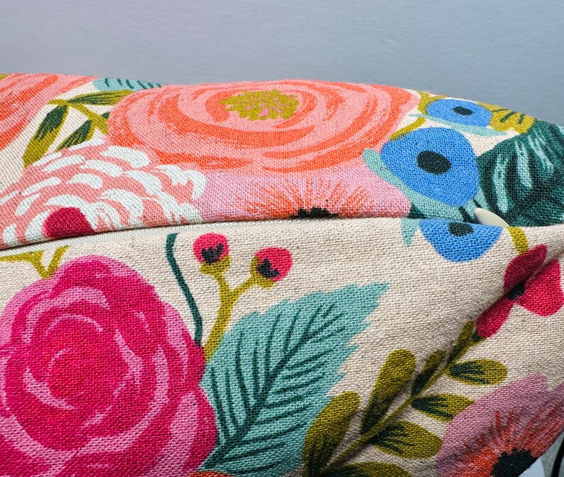 May include: Close-up of a floral patterned pillow. The pillow features large, colorful flowers in shades of pink, orange, and blue, set against a beige background. Green leaves and stems complete the design, creating a vibrant, botanical theme.