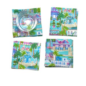 Puede incluir: Cuatro posavasos cuadrados de tela turquesa con una escena tropical con fuentes, palmeras y bicicletas. Un posavasos tiene un vaso transparente encima. Los posavasos son una adición vibrante a cualquier decoración del hogar.