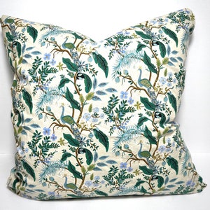 May include: Cream-colored decorative pillow with a repeating pattern of peacocks, foliage, and flowers in shades of green, blue, and brown. The pillow has a soft, textured appearance and is likely made of fabric. The design gives a botanical and nature-inspired theme.
